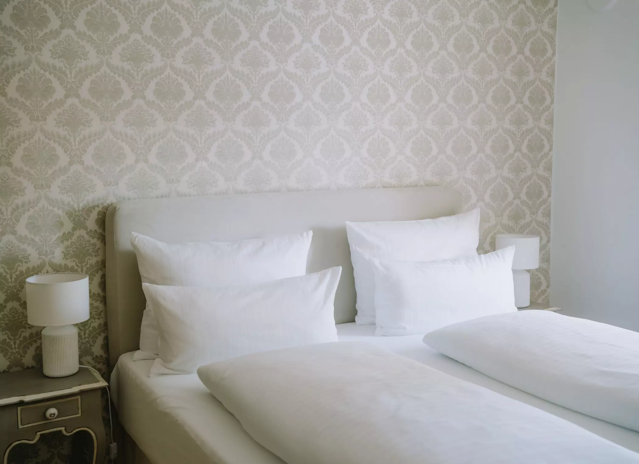 Helles Hotelzimmer mit Doppelbett, weißer Bettwäsche und Nachttisch mit Lampe.